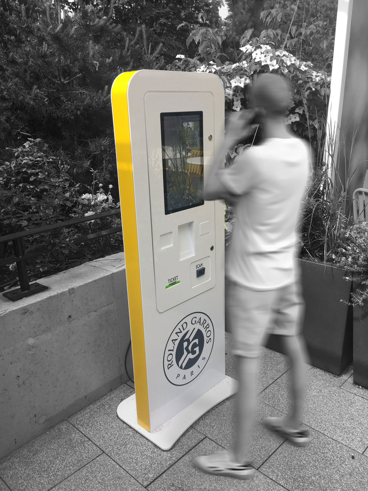 Une personne interagit avec un kiosque en libre-service de Roland Garros à l'extérieur, entouré de verdure, l'image en couleur sélective mettant en évidence le kiosque.