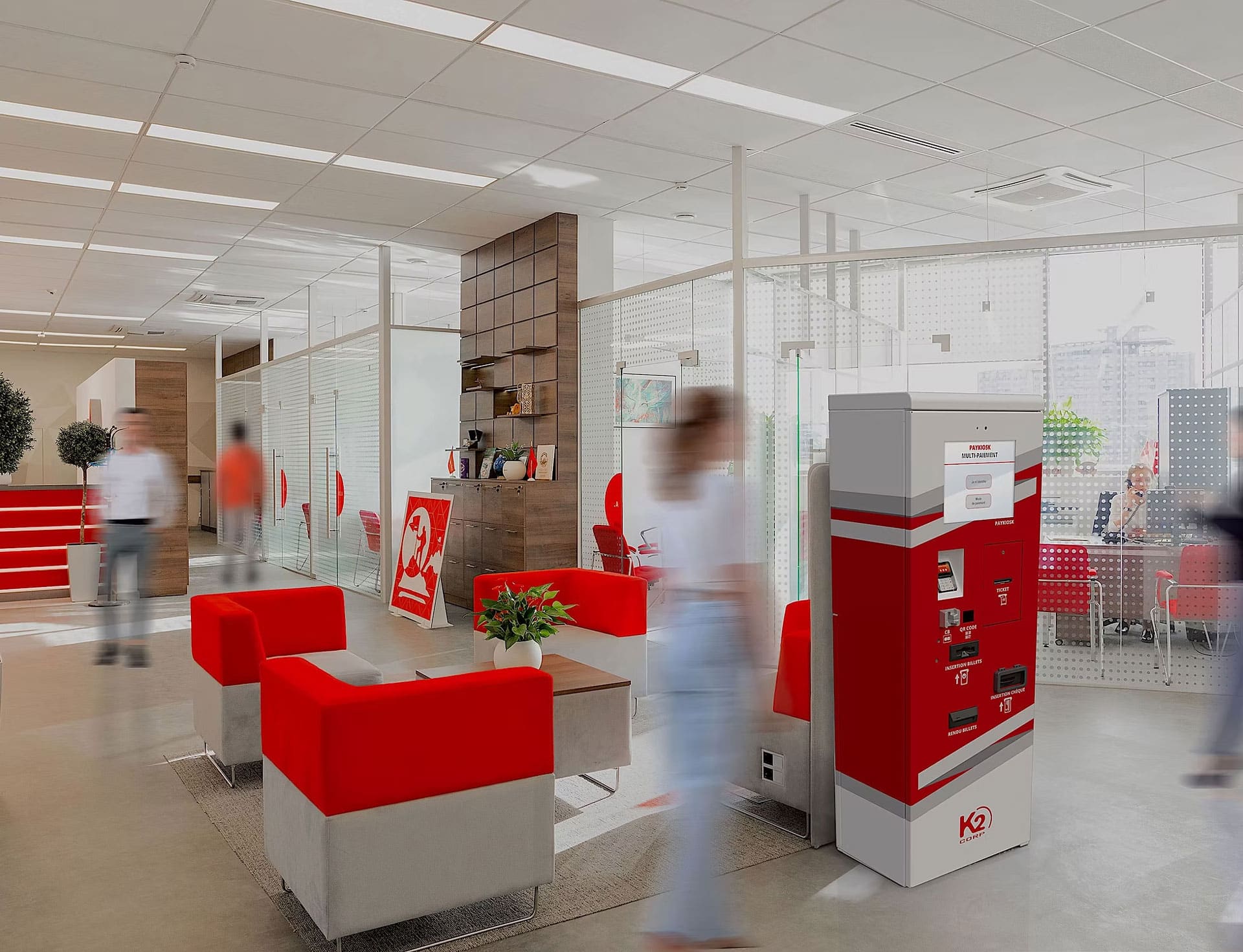 Hall d'entrée de bureau moderne avec des sièges rouges et gris, un kiosque libre-service, des salles de réunion aux parois vitrées et des personnes marchant et assises en arrière-plan.