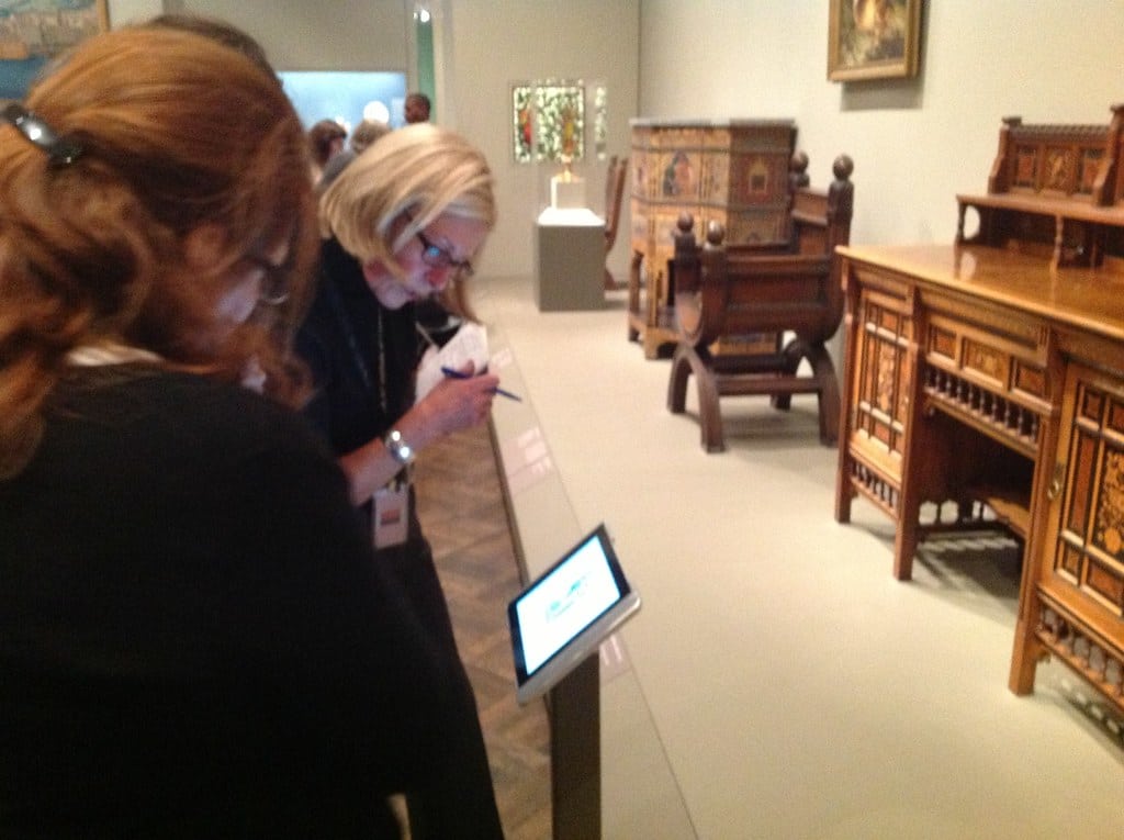 Deux femmes se tiennent dans une galerie de musée, l'une écrivant sur un bloc-notes et l'autre utilisant un totem tactile devant des meubles en bois ornés.